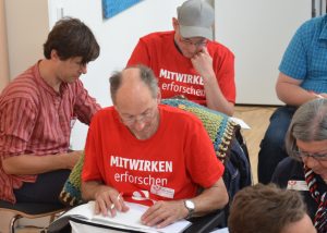 Auf der Jahrestagung 2018 im Plenum: Teilnehmende sind damit beschäftigt, etwas aufzuschreiben. Zwei Männer tragen ein rotes T-Shirt mit dem Aufdruck "MITWIRKEN erforschen".