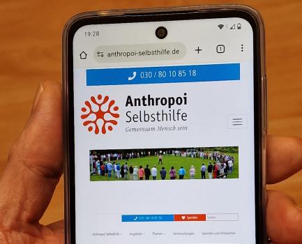 Smartphone in einer Hand. Man sieht darauf einen Ausschnitt der Website von Anthropoi Selbsthilfe.